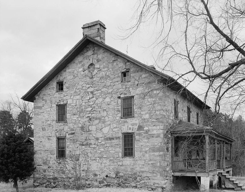 Black and white photograph of a two-storey stone house with a gabled roof and central chimney, bare trees overhead and a covered porch structure at the right-hand end.
