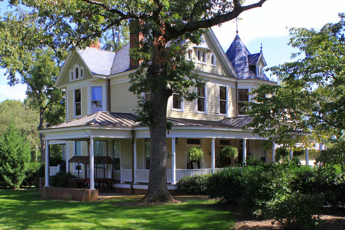 Two-storey Queen Anne house painted cream and grey, with a wraparound porch, a conical turret roof and a large oak tree on the front lawn.