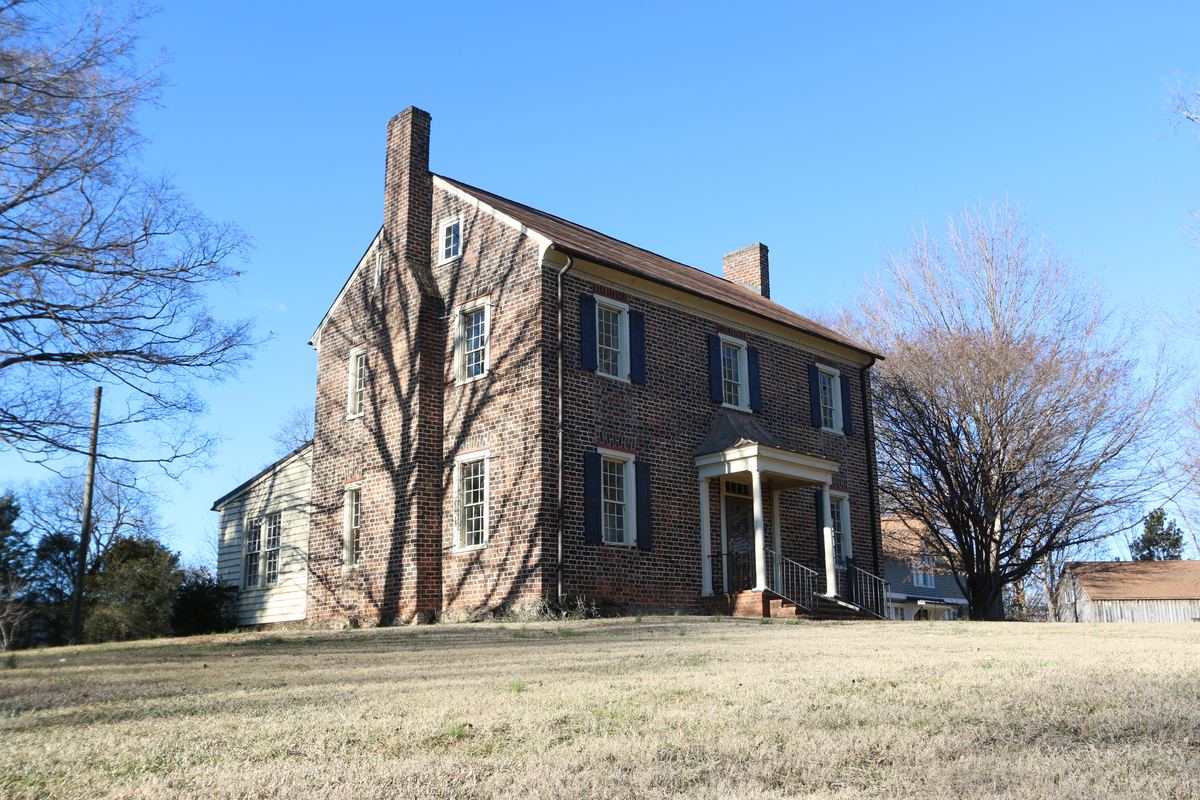 Two-storey brick house with a tall exterior end chimney and a small columned entrance porch, standing on a rise above a dry lawn.