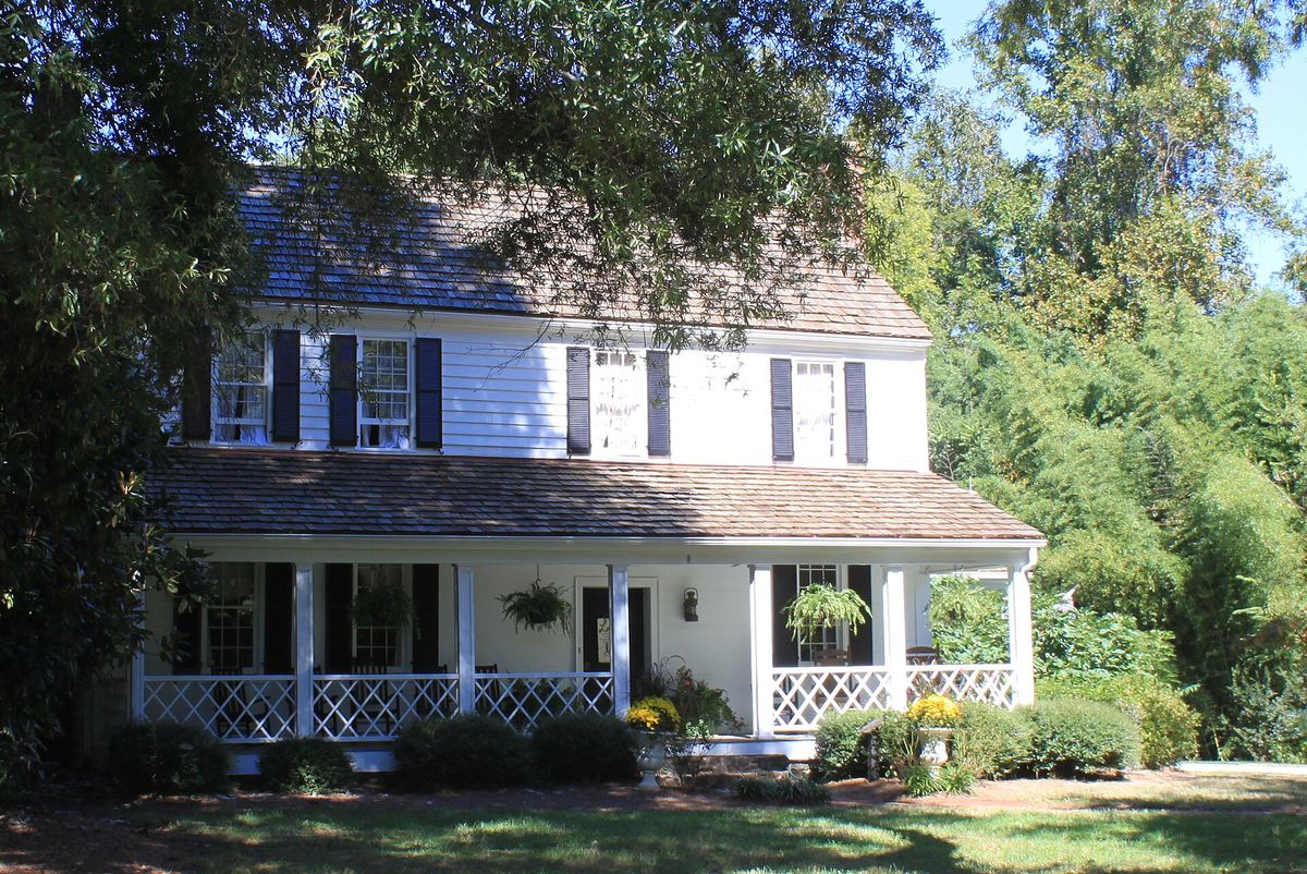 White clapboard house with dark shutters, a full-width covered porch and a steep shingled roof, set among mature trees.