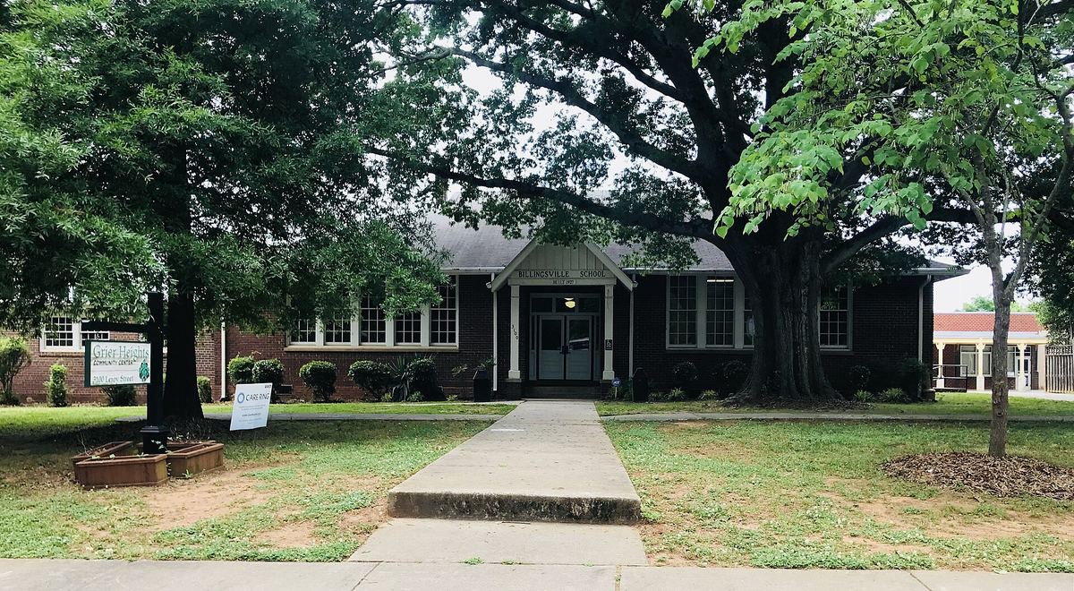 Low single-storey brick school building with a central entrance, approached by a straight concrete path between two very large mature trees.