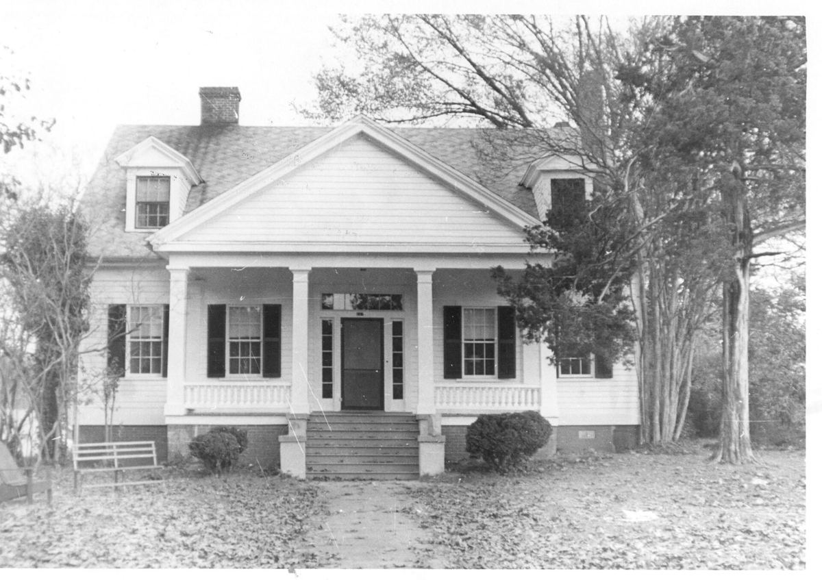 Black and white photograph of a white Greek Revival house with four tall square columns supporting a full-height portico, surrounded by bare trees.