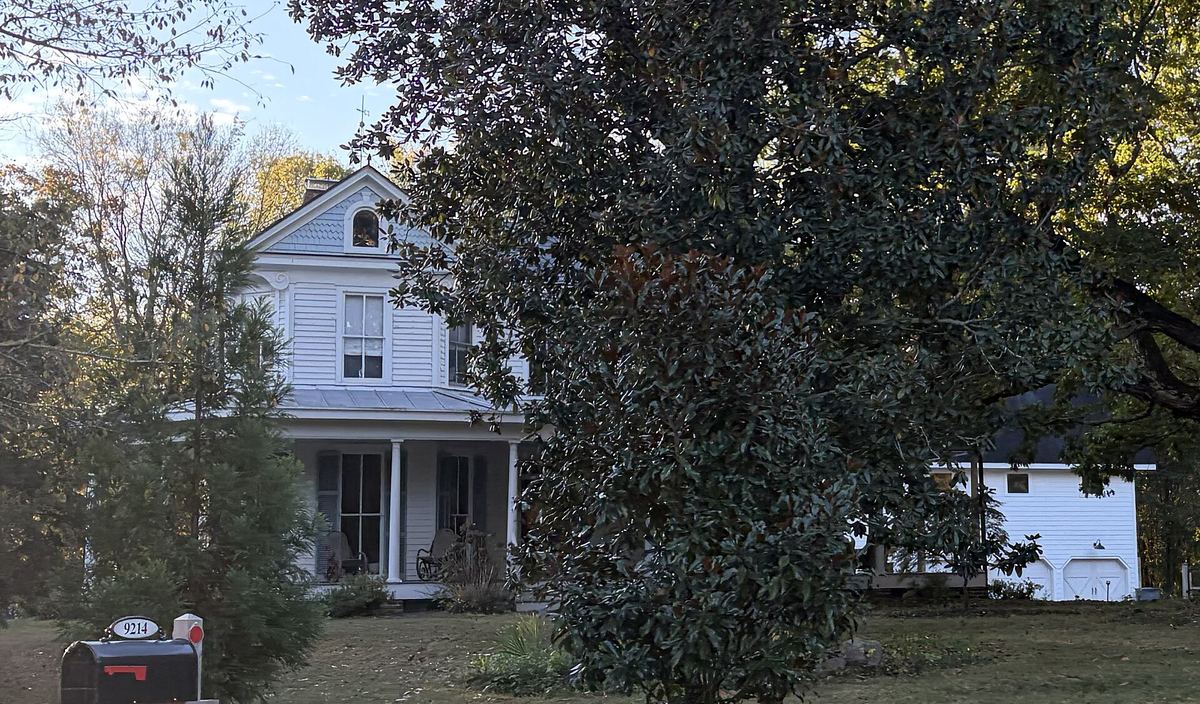 White two-storey farmhouse with a covered porch, largely screened by a large dark evergreen tree in the foreground.