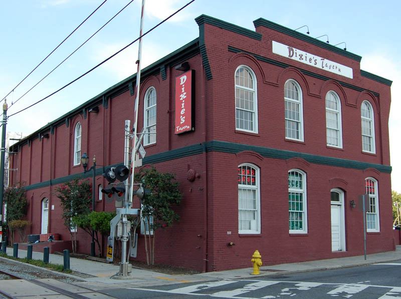 Two-storey dark red brick commercial building on a street corner, with tall arched windows picked out in white, a painted green band at first-floor level, and a hanging sign and fascia board reading 'Dixie's Tavern'. A level-crossing signal and rails cross the foreground.