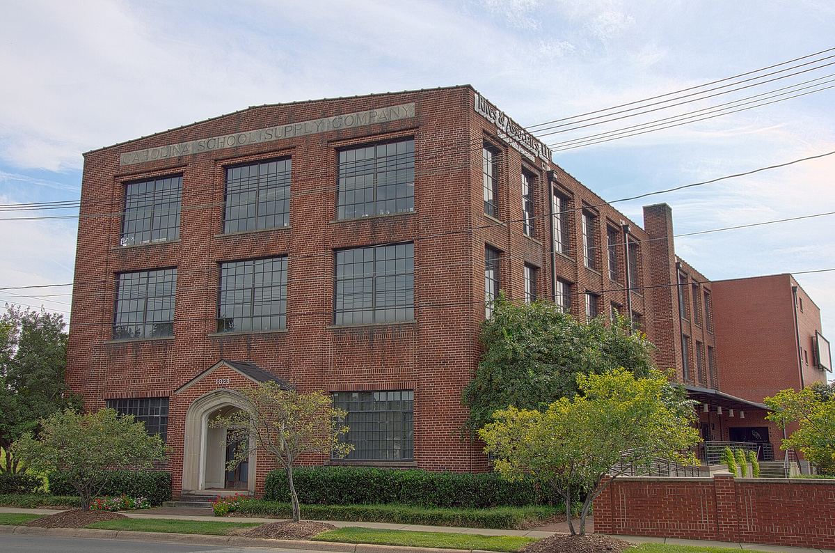 Three-storey red brick commercial building with regular banded windows and an arched stone entrance surround, set behind low shrubs.