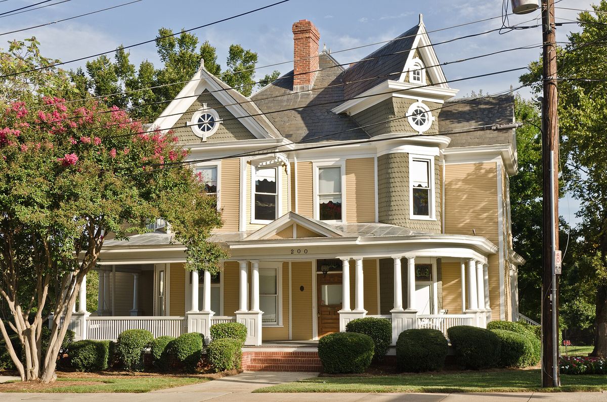 Two-storey house with cream shingled gables, decorative bargeboards and a bay-fronted porch, behind flowering crape myrtles.