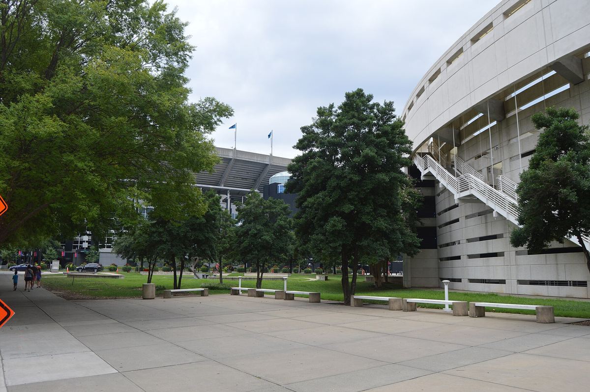 View across a paved plaza and lawn towards a large curving concrete stadium structure with external staircases.