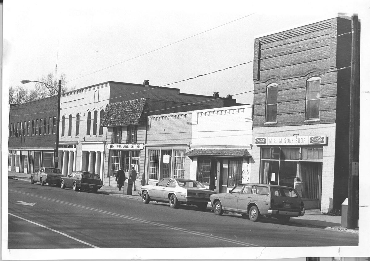 Black and white photograph of a row of two-storey brick commercial buildings along a main street, with 1980s cars parked at an angle at the kerb.