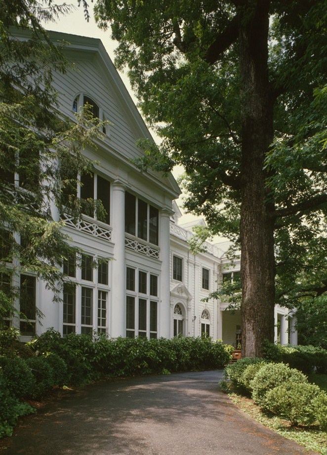 White painted house with a two-storey columned portico and decorative balustrade, glimpsed through the trunks of very large trees along a driveway.