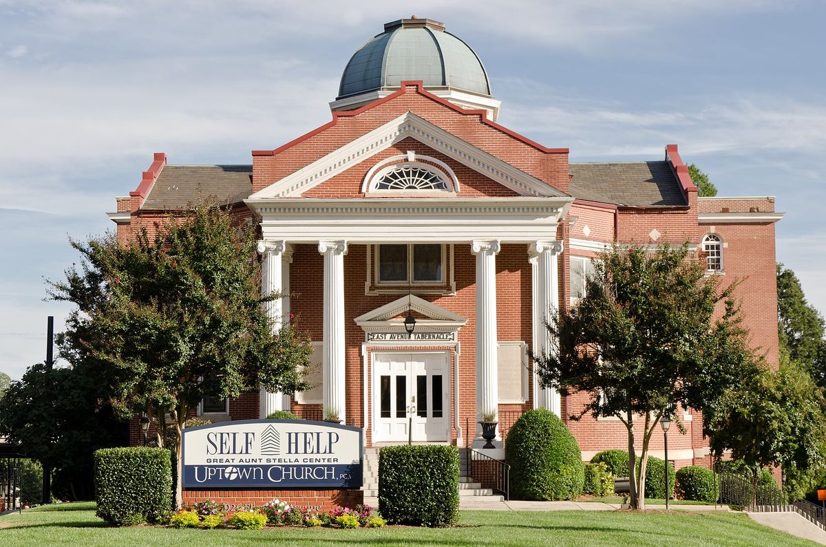 Red brick church with a domed cupola, a white columned portico and a sign reading Self Help and Uptown Church on the lawn in front.