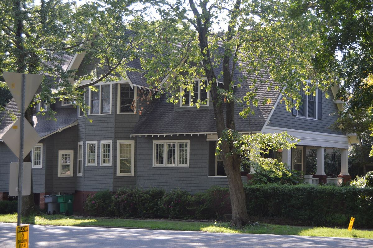 Grey shingled house with white trim and a deep front porch, partly screened by a large tree, on a residential street.