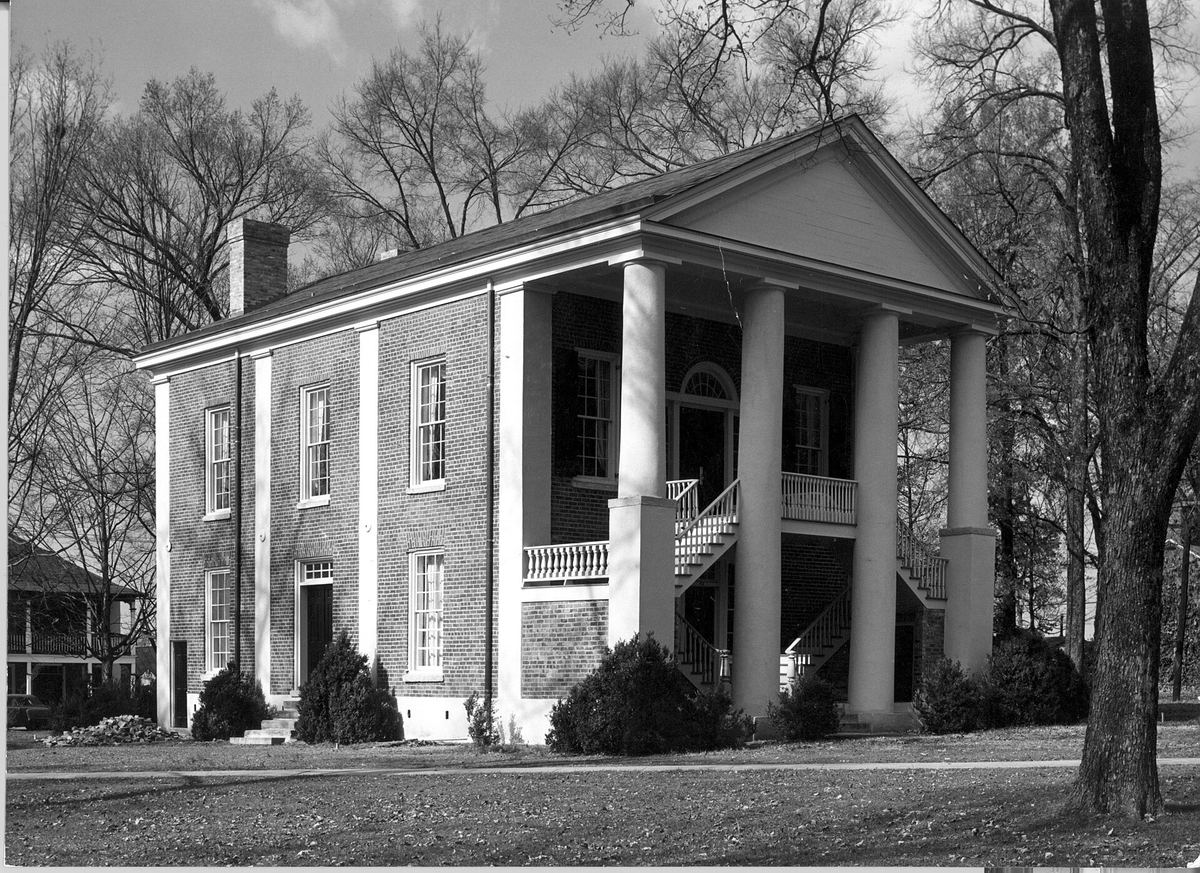 Black and white photograph of a two-storey brick building with a full-height portico of four slender columns, set among bare trees.