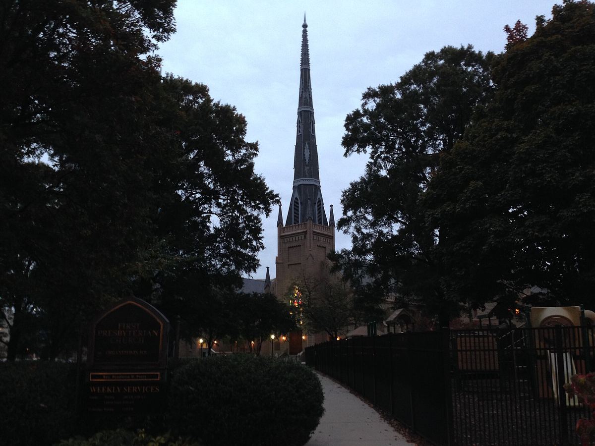 Dark evening view of a tall church spire rising above trees, with a lit path and railings in the foreground.