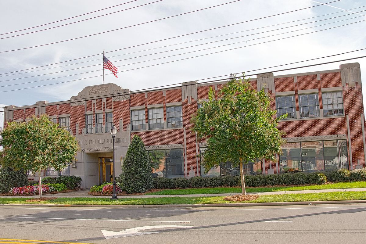 Long two-storey brick industrial building with tall steel-framed windows, a flag on a pole above the entrance and a wide road in front.