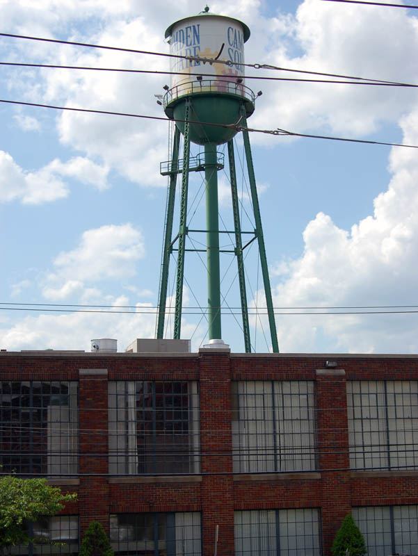 Green steel water tower on tall lattice legs rising above a red brick mill building.