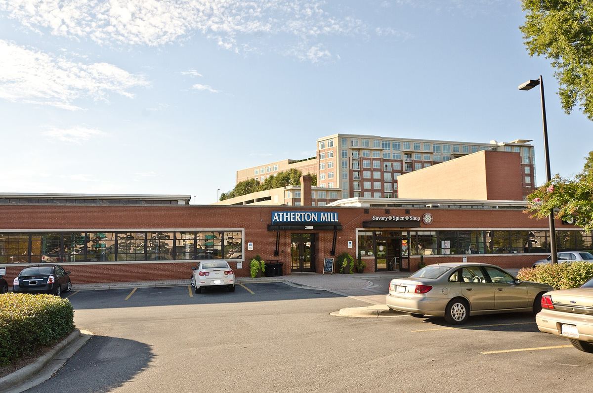 Low single-storey retail building with a sign reading Atherton Mill, cars parked in front and taller modern blocks behind.