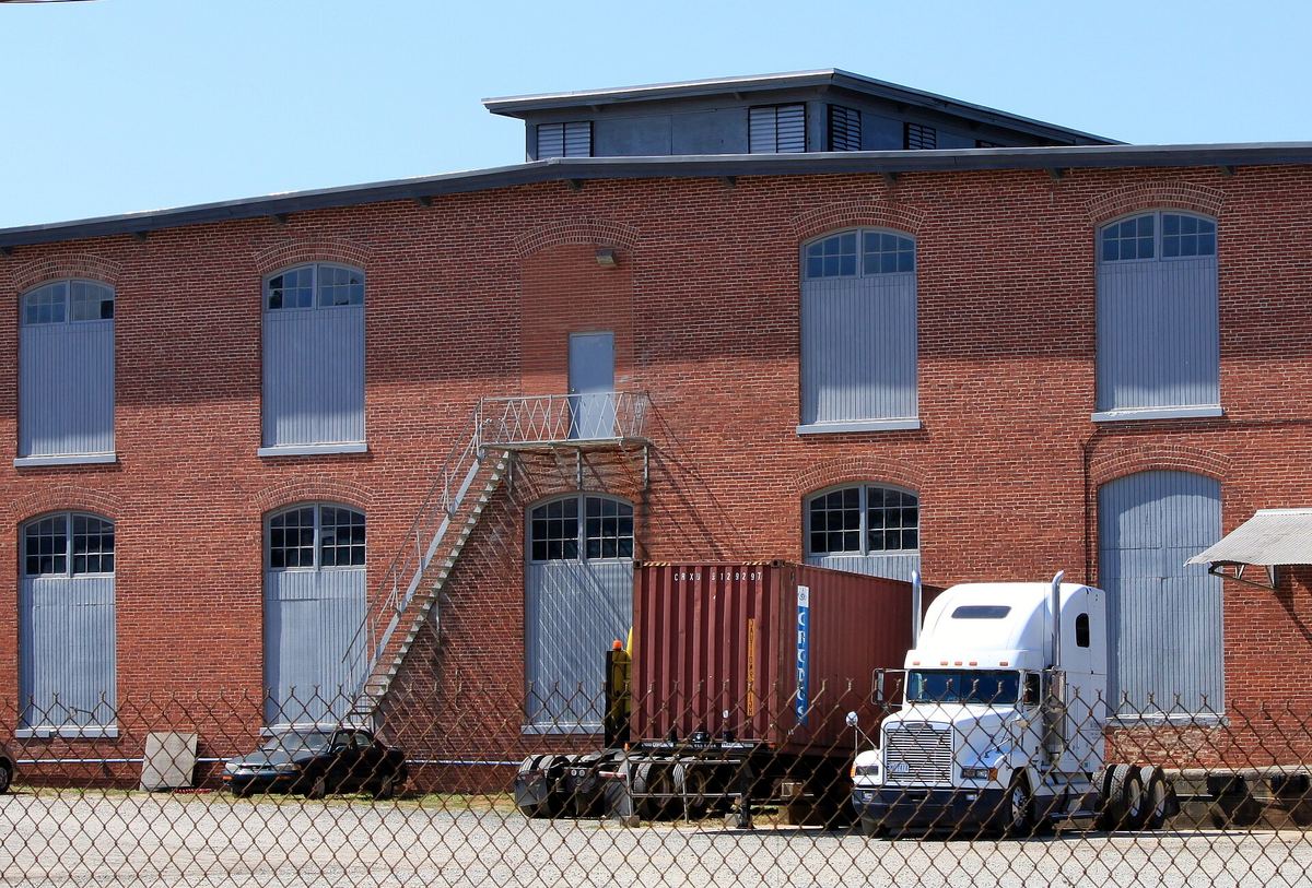 Two-storey red brick mill building with tall arched windows and an external steel staircase, with a lorry and shipping container parked behind a chain-link fence.