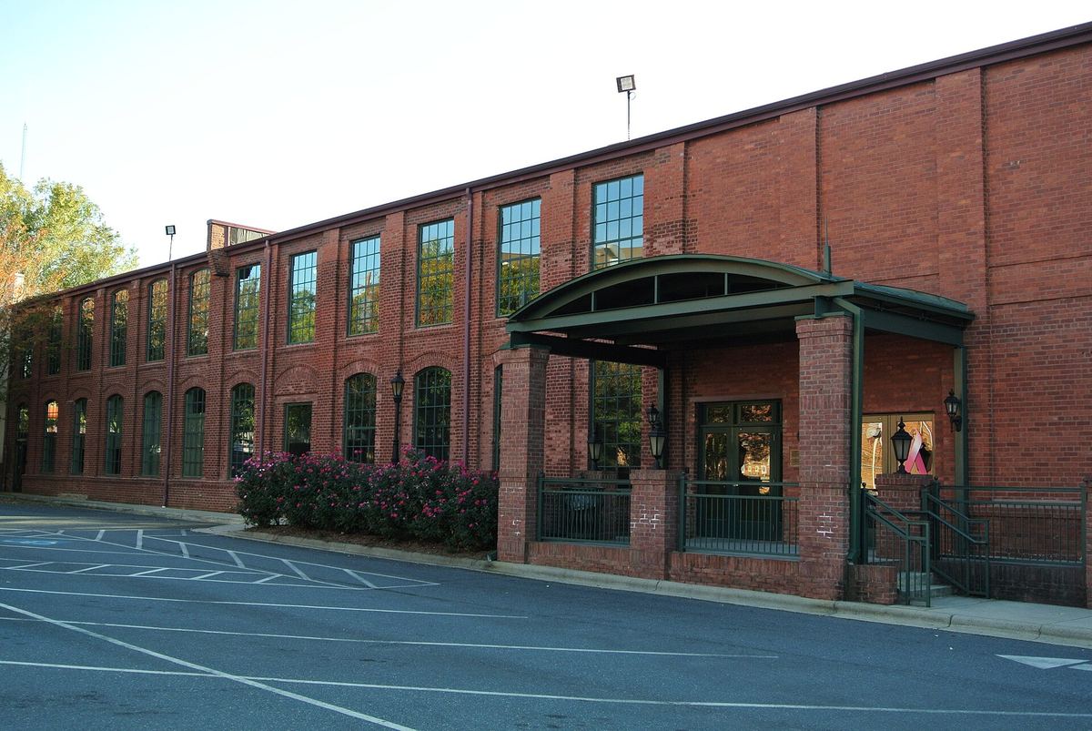 Long low red brick industrial building with a continuous row of arched windows and a green canopied entrance, fronted by an empty car park.