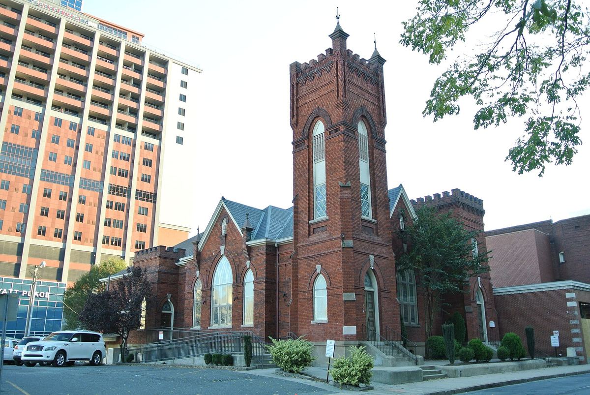 Red brick church with a tall crenellated corner tower and pointed arched windows, with modern high-rise blocks behind.