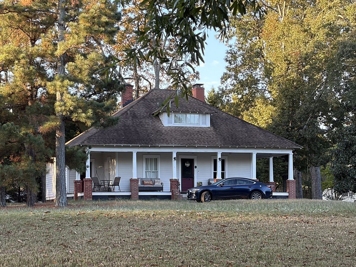 Single-storey bungalow with a deep porch on brick piers and a hipped roof, on a large open lawn with mature trees behind.