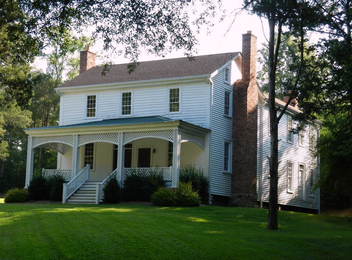 White weatherboarded two-storey house with a wide covered porch and a tall brick chimney at the gable end, on a mown lawn.