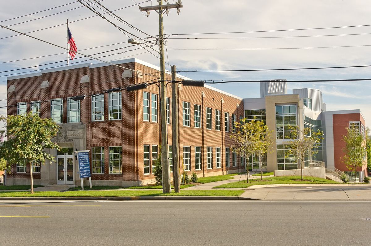 Two-storey brick industrial building on a corner with a flagpole, adjoining a lower modern extension, with power lines overhead.