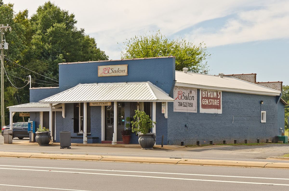 Single-storey grey painted store building with a metal awning, hanging sign and planters, directly on the roadside.