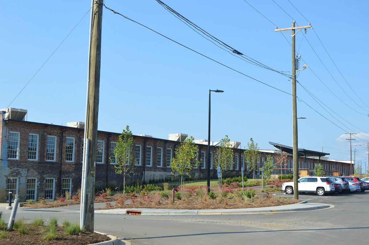 Long low brick mill building with regular windows, seen across a wide road junction with young trees and overhead power lines.