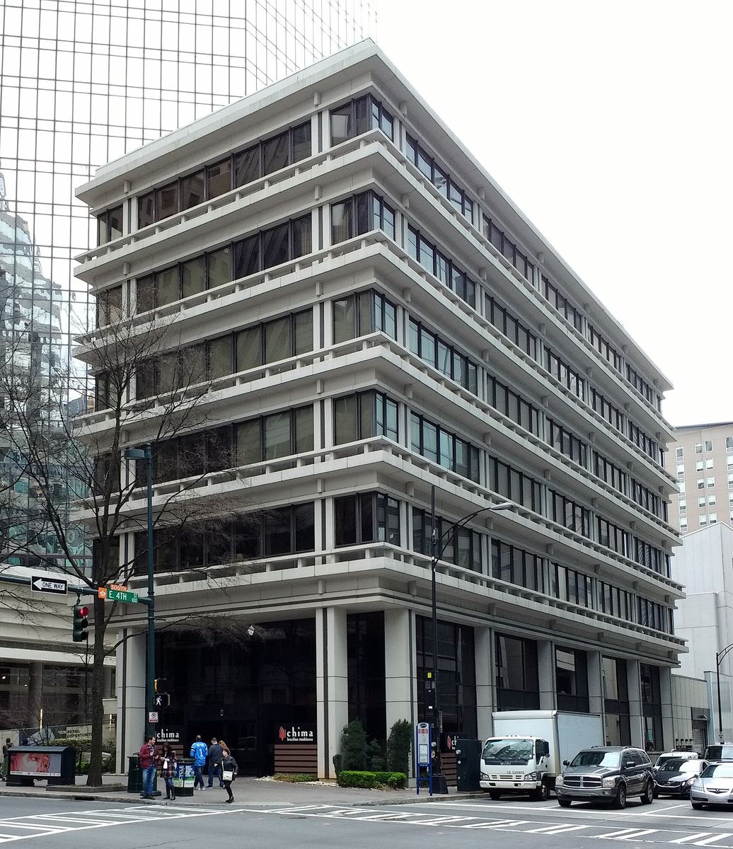 Six-storey mid-century office building with strongly projecting horizontal floor bands, on a street corner with a mirrored tower rising behind.