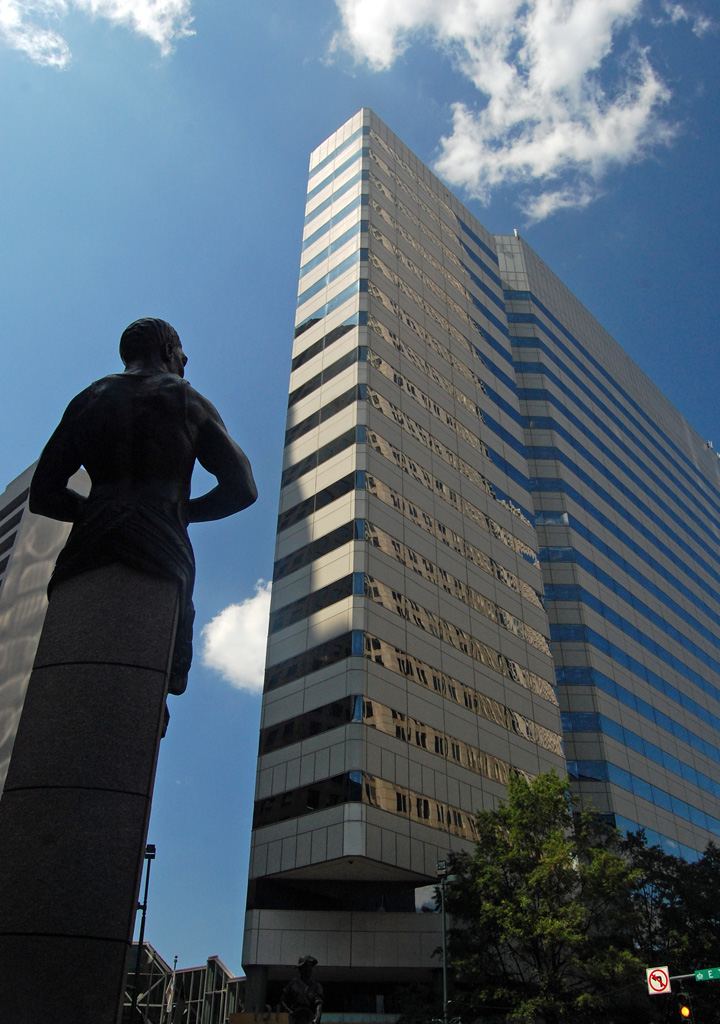Looking up at a tall modern office tower clad in blue and silver glass, with a dark bronze statue of a standing figure in the foreground.