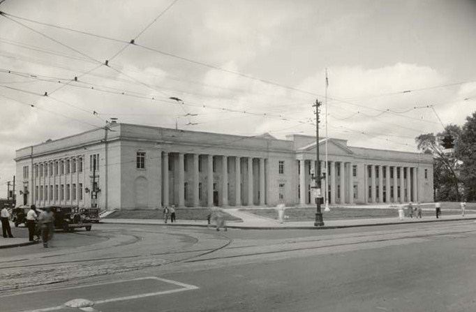 Black and white photograph of a long neoclassical stone federal building with a colonnade of square columns, tram wires overhead and a near-empty street in front.