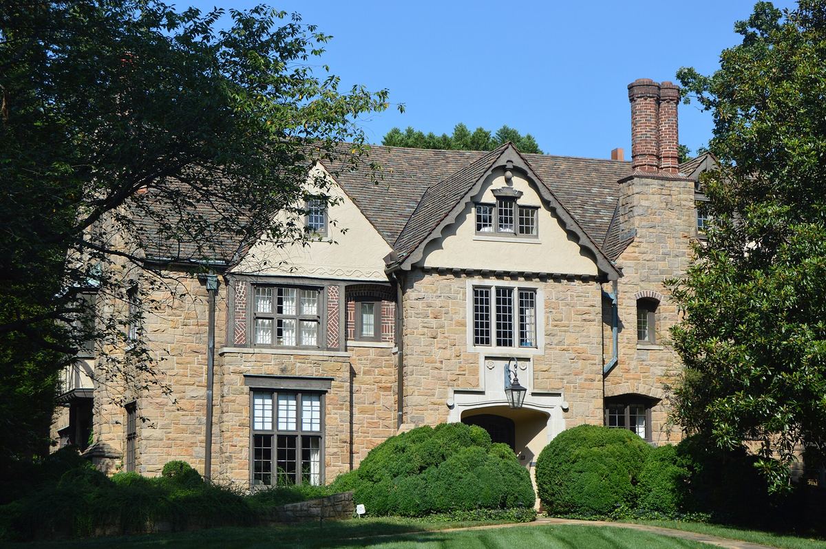 Large stone Tudor Revival house with a steeply pitched roof, tall chimney stack and leaded windows, behind clipped hedges.