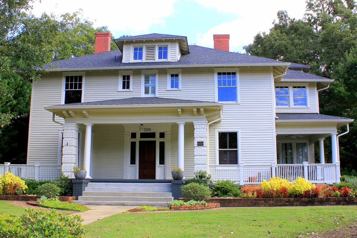 Pale blue and white four-square house with a columned entrance porch, dormer window and two red brick chimneys, with flower beds in front.
