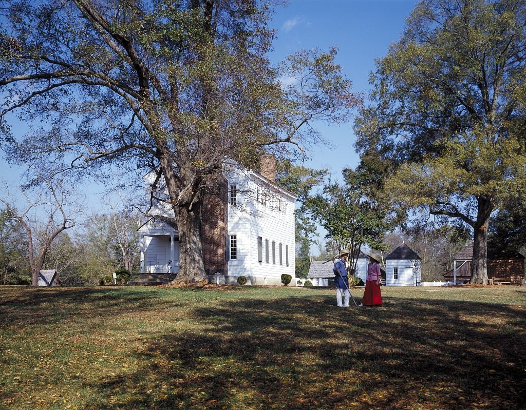 White clapboard plantation house seen across a wide lawn beneath a very large bare tree, with two people in period costume standing in the middle distance.