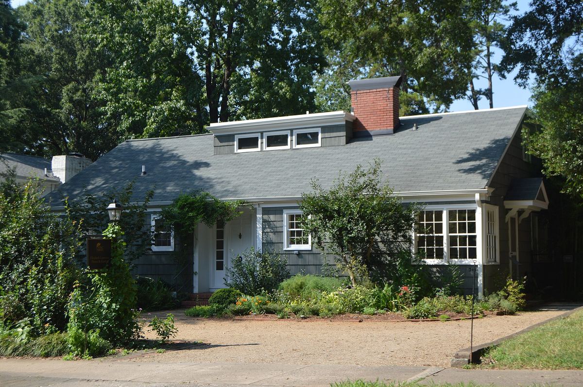 Single-storey white painted house with a low-pitched roof and a brick chimney, partly screened by dense shrubs and planting.
