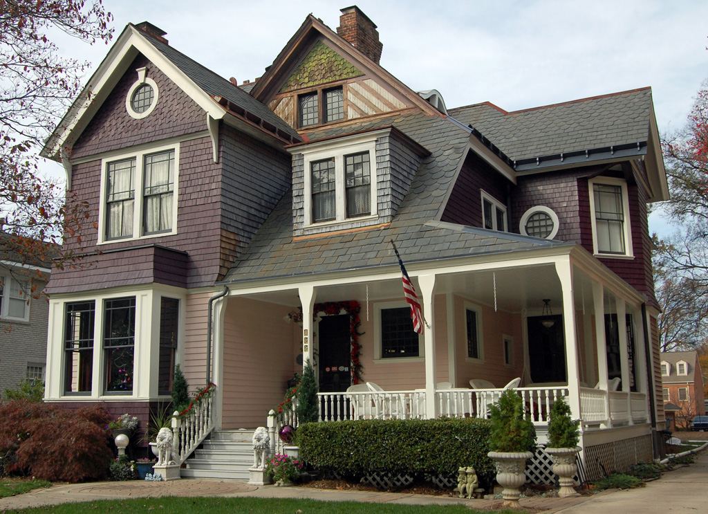 Two-and-a-half storey shingled Queen Anne house painted mauve and cream, with a wraparound porch, decorative gables and an American flag by the door.