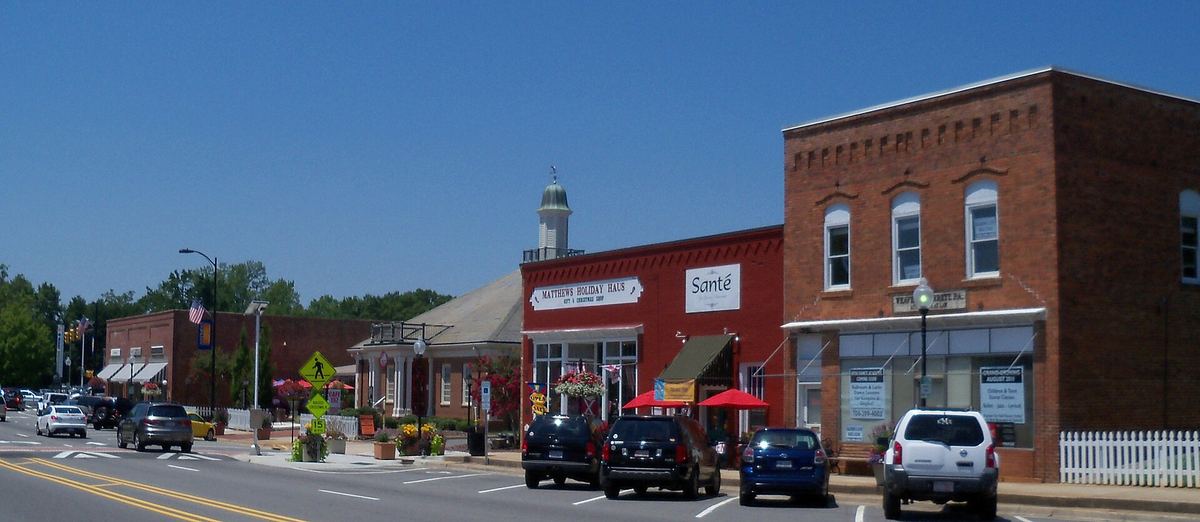 Small-town main street with two-storey brick commercial buildings, shopfronts with awnings, parked cars and a clock on a post.