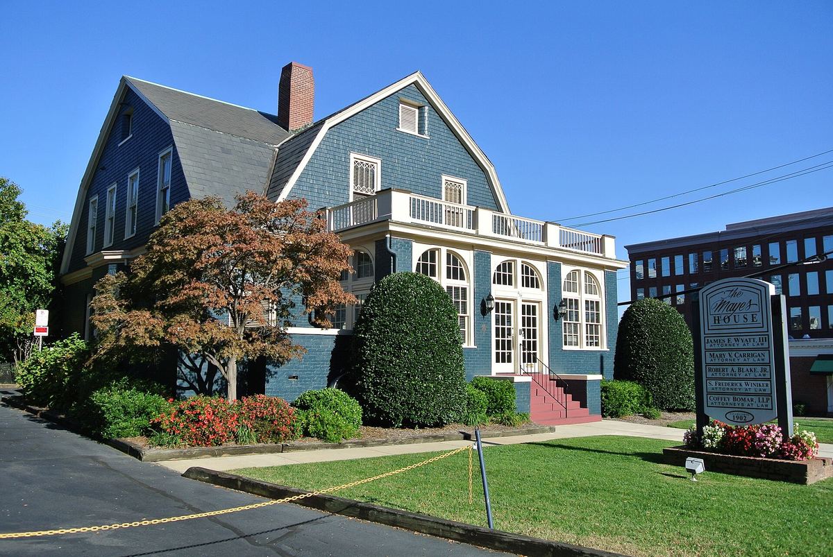 Large dark green shingled house with white trim, a two-storey bay of windows and a clipped conical shrub on the front lawn beside a pavement.