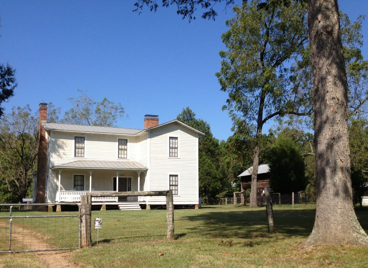 White clapboard farmhouse with a covered porch, seen across a fenced paddock with a very large mature tree in the foreground.