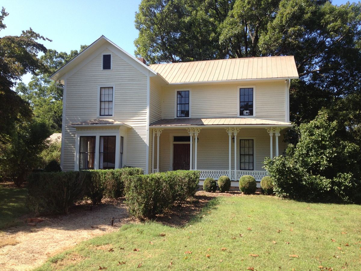 Two-storey cream weatherboarded farmhouse with a metal roof, gabled wing and a covered porch, on a sloping lawn.