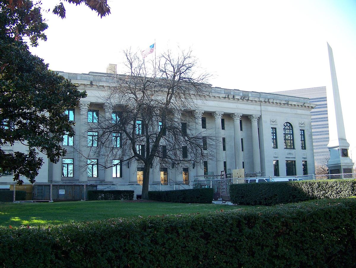 Large pale neoclassical courthouse with a colonnade of tall columns, behind a hedge and bare tree, with a thin obelisk visible to the right.