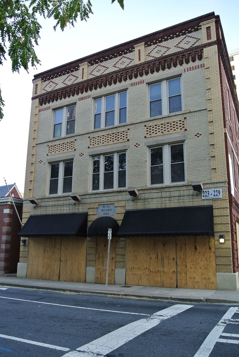 Three-storey pale brick commercial building with decorative patterned brickwork at the cornice, black awnings and boarded ground-floor openings.