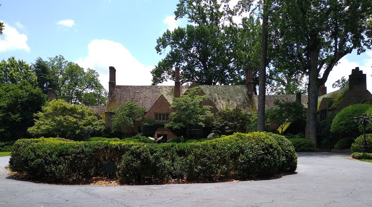 Large house with steeply pitched rooflines and tall chimneys, largely hidden behind a very high clipped hedge along a driveway.