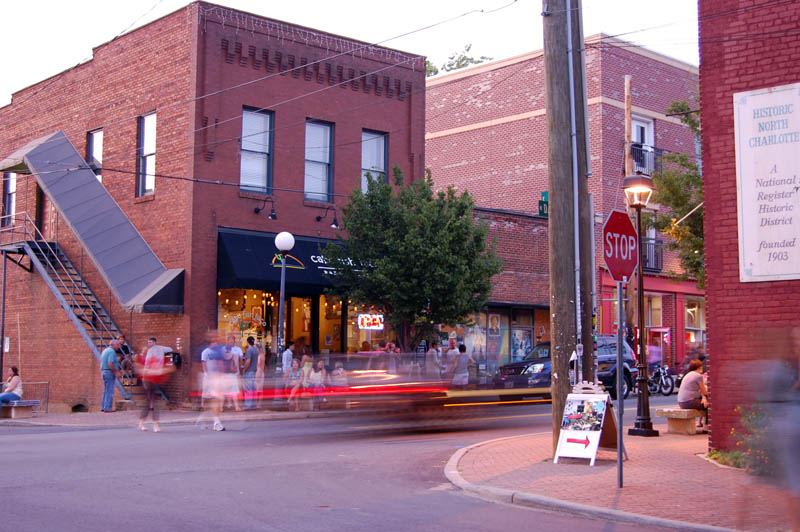 Evening street scene of red brick two-storey commercial buildings with lit shopfronts, a stop sign and blurred movement of people and traffic.