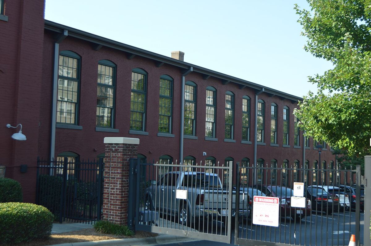 Long two-storey red brick mill building with a regular row of windows, behind a black metal fence with cars parked alongside.