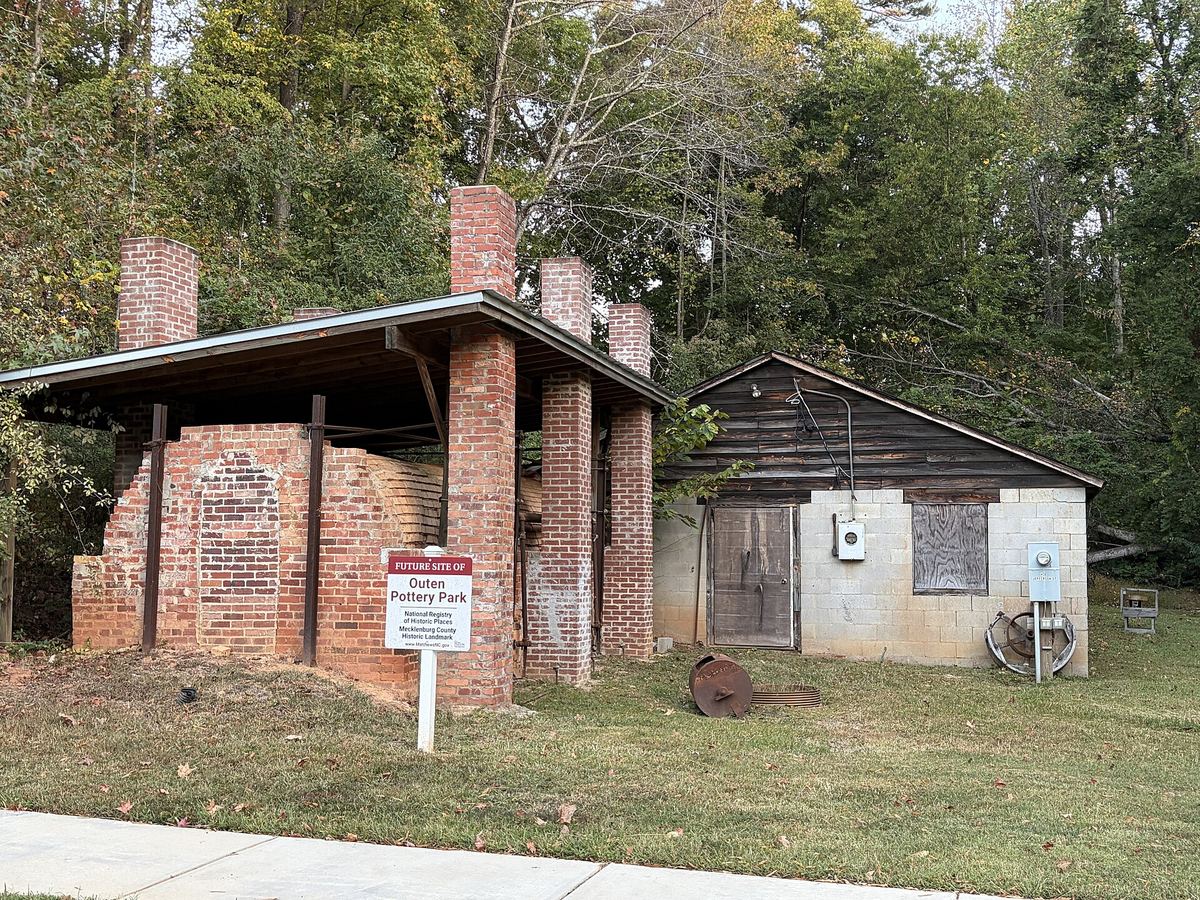 Open-sided brick kiln structure and a small rendered outbuilding on a grassy site, with an interpretive sign in the foreground.