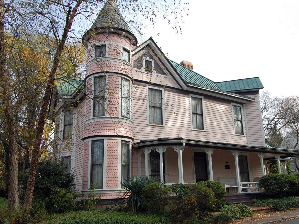 Two-and-a-half storey Victorian house with a round corner turret, green metal roof and wraparound porch, among bare autumn trees.