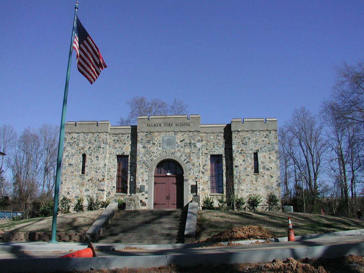 Single-storey building of rough multicoloured fieldstone with a crenellated parapet, a round-arched entrance with red double doors reached by a flight of steps, and the words 'PALMER FIRE SCHOOL' carved into the stone lintel above. An American flag flies from a pole at the left; the foreground is an unfinished kerb and bare earth.