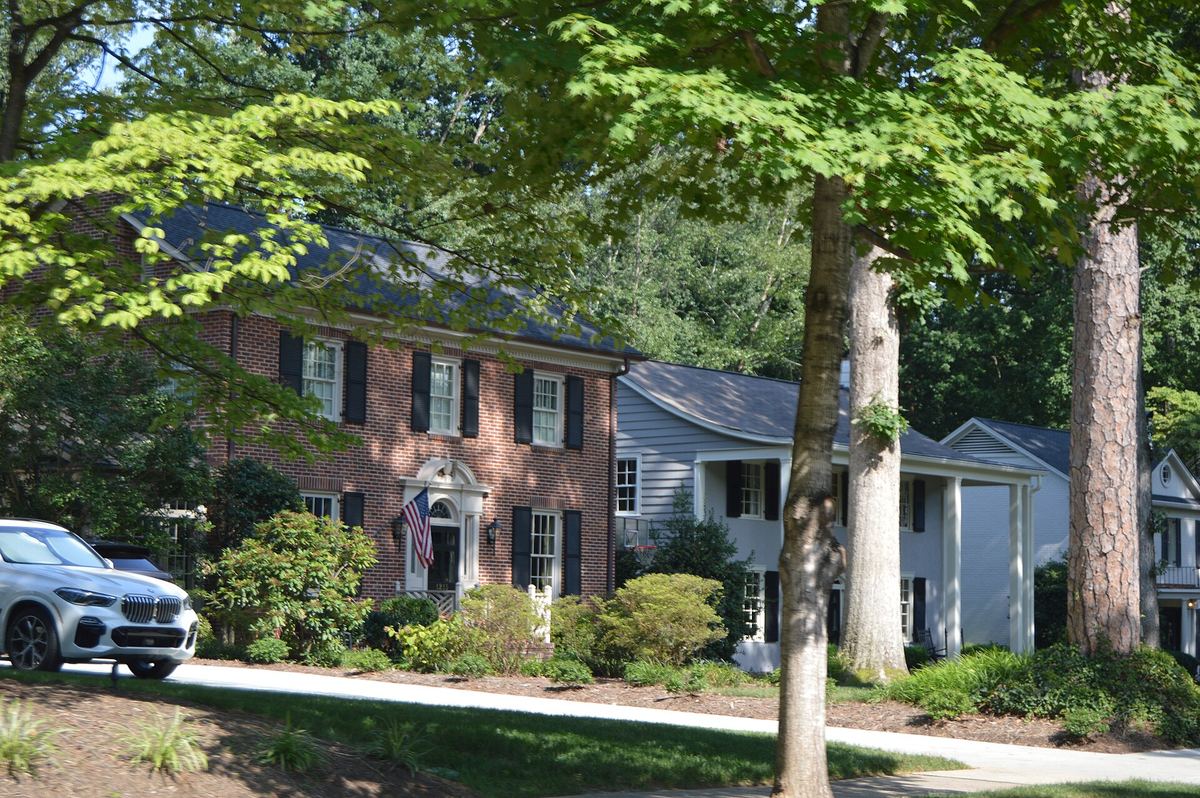 Two-storey brick house with white-framed windows and a flag beside the door, on a tree-lined residential street with a parked car.