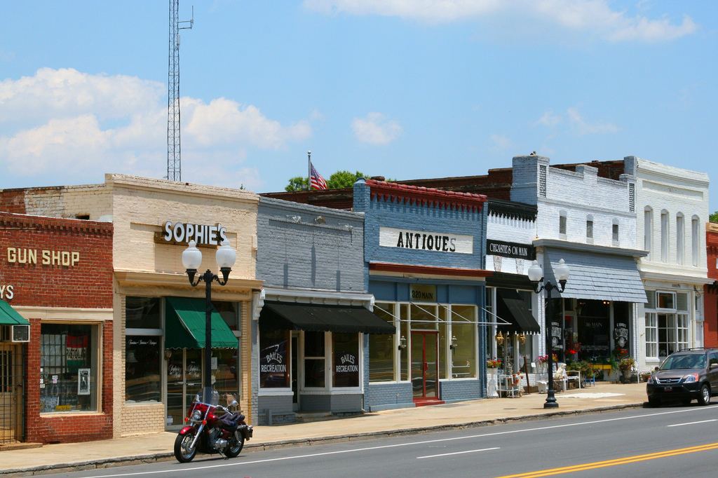 Row of one and two-storey small-town shopfronts with signs reading Gun Shop, Sophie's and Antiques, a motorcycle parked at the kerb.